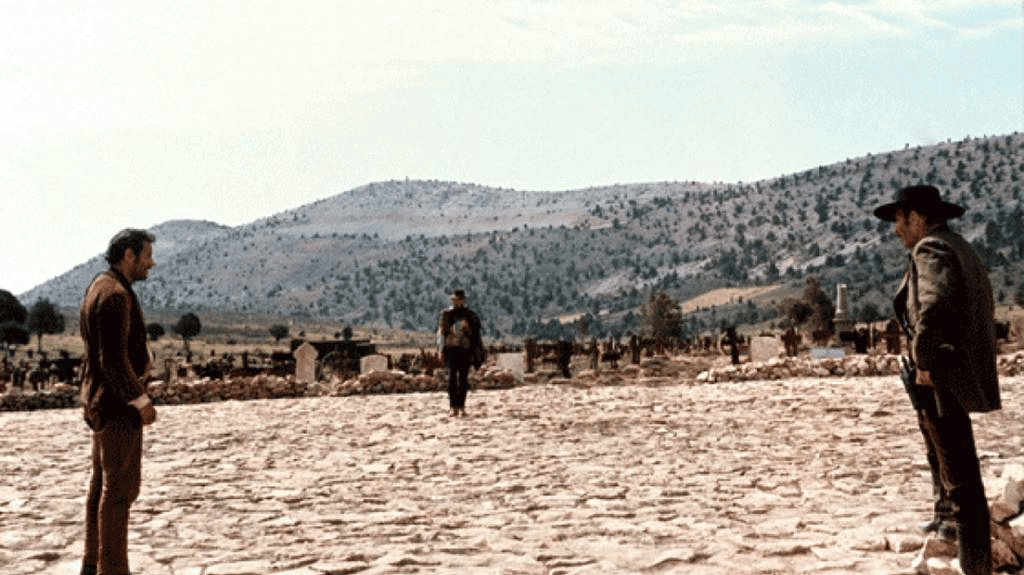 Three men facing each other in a dusty setting with mountains in the background, suggesting a tense standoff.
